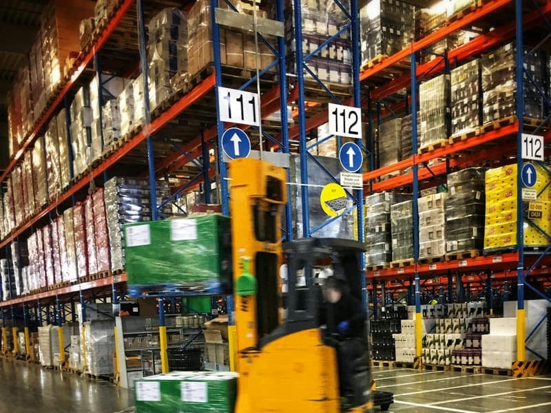 A forklift carrying a loaded pallet down an aisle of a distribution warehouse, with racking stacked to the ceiling on both sides.